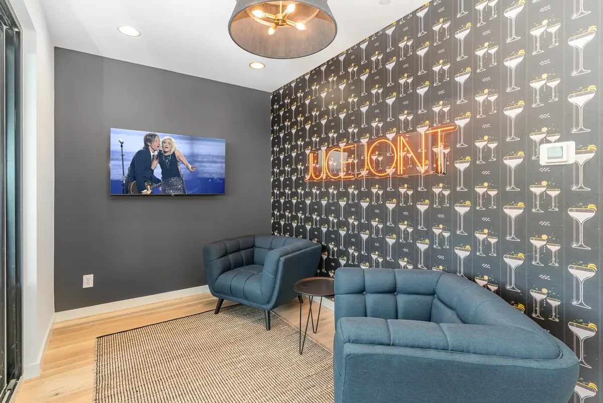 A playful and inviting interior lounge area highlighted by blue armchairs, a neon sign, a wall-mounted television, and bold patterned wallpaper. The arrangement encourages conversation and entertainment, ideal for relaxing or watching live performances.