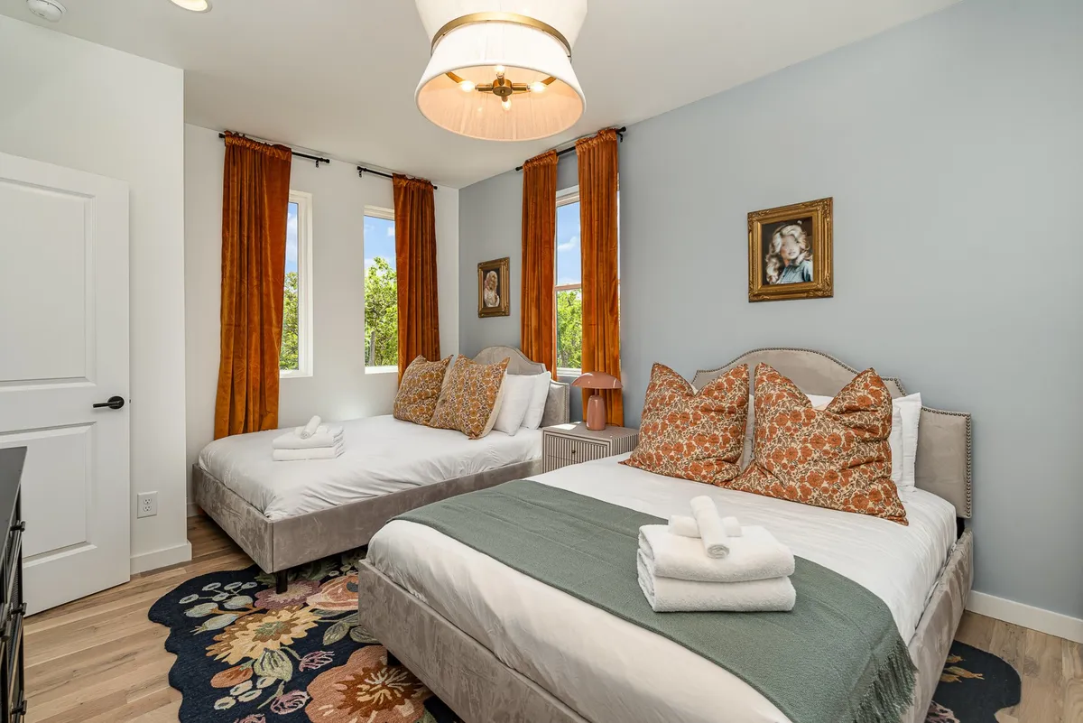 A bright, welcoming bedroom featuring two beds with decorative pillows, a vibrant area rug, large windows letting in natural light, and tasteful framed artwork on the wall. An elegant light fixture adds a refined touch to the cozy atmosphere.
