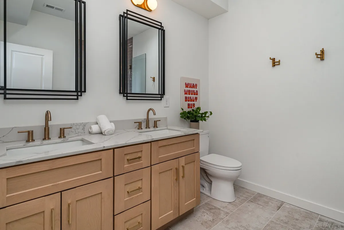 A cheerful double vanity bathroom with large mirrors, gold faucets, decorative wall art, and a potted plant. The bright and inviting design promotes both function and style, ideal for shared routines.