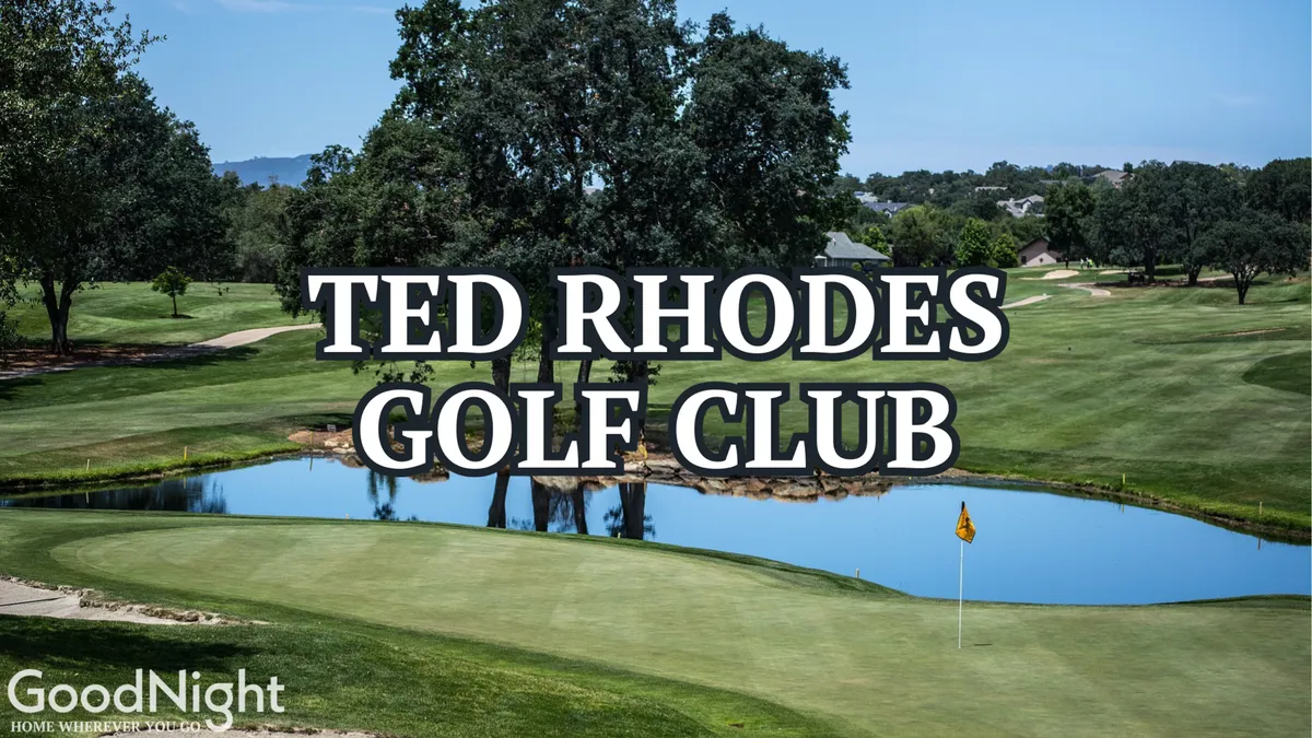 A picturesque view of the Ted Rhodes Golf Club, showing an expansive green course bordered by a pond and mature trees, with a flagstick visible. The tranquil scene invites both socializing and friendly competition in a scenic setting.