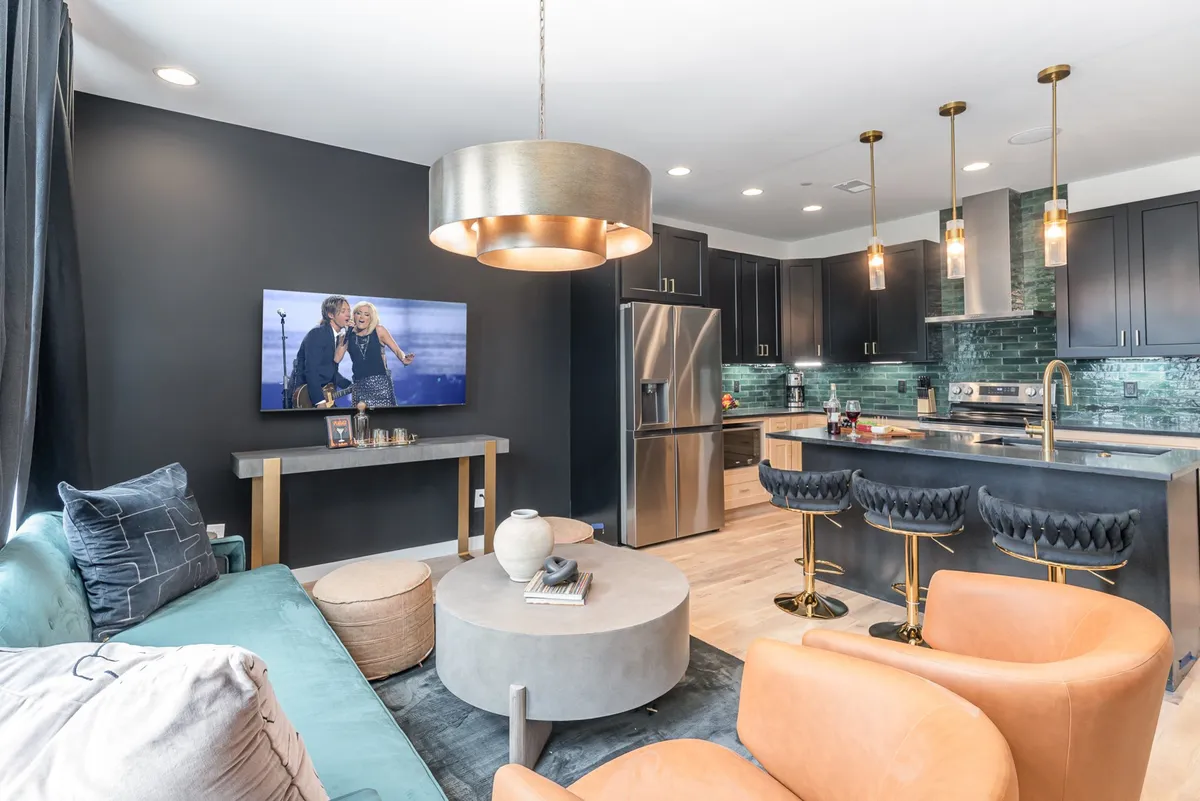 A contemporary open-concept living area featuring a chic sofa, coffee table, and a modern kitchen island in view. The space is well-lit, with stylish lighting fixtures and a large television, creating an atmosphere ideal for social gatherings, movie nights, or relaxed lounging.