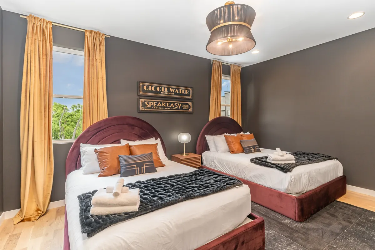 A comfortable and stylish bedroom with two beds featuring plush headboards, a vintage-style sign on the wall, warm-toned curtains, and a chic overhead light fixture. The setup feels inviting for both relaxation and socializing.