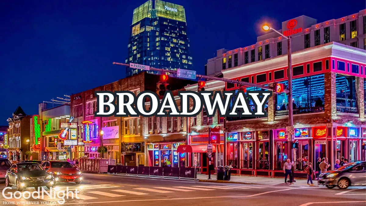 A vibrant exterior nighttime scene featuring neon signs, a bustling street full of people walking, cars, and a variety of buildings. The energetic atmosphere and visible activity suggest a lively nightlife district, reminiscent of Nashville’s Broadway.