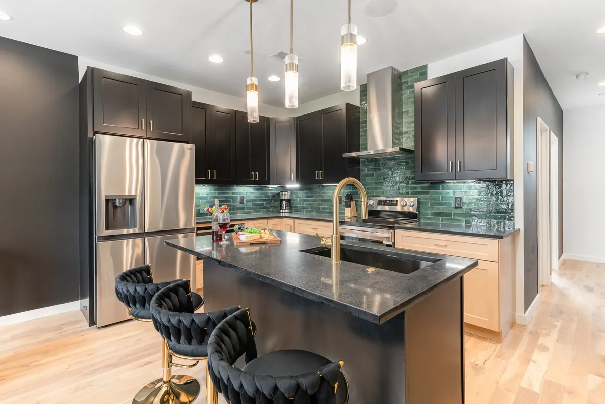 This image showcases a modern kitchen with a black kitchen island, gold pendant lights, a green backsplash, and stainless steel appliances. The space is clearly designed for cooking and entertaining, with inviting textures and finishes.